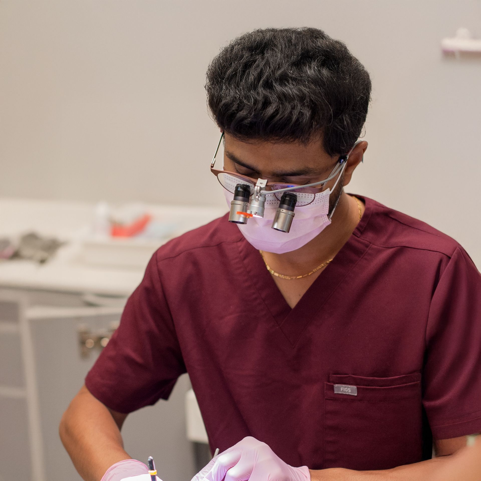 Dentist in burgundy scrubs, mask, and magnification glasses working in a dental office.