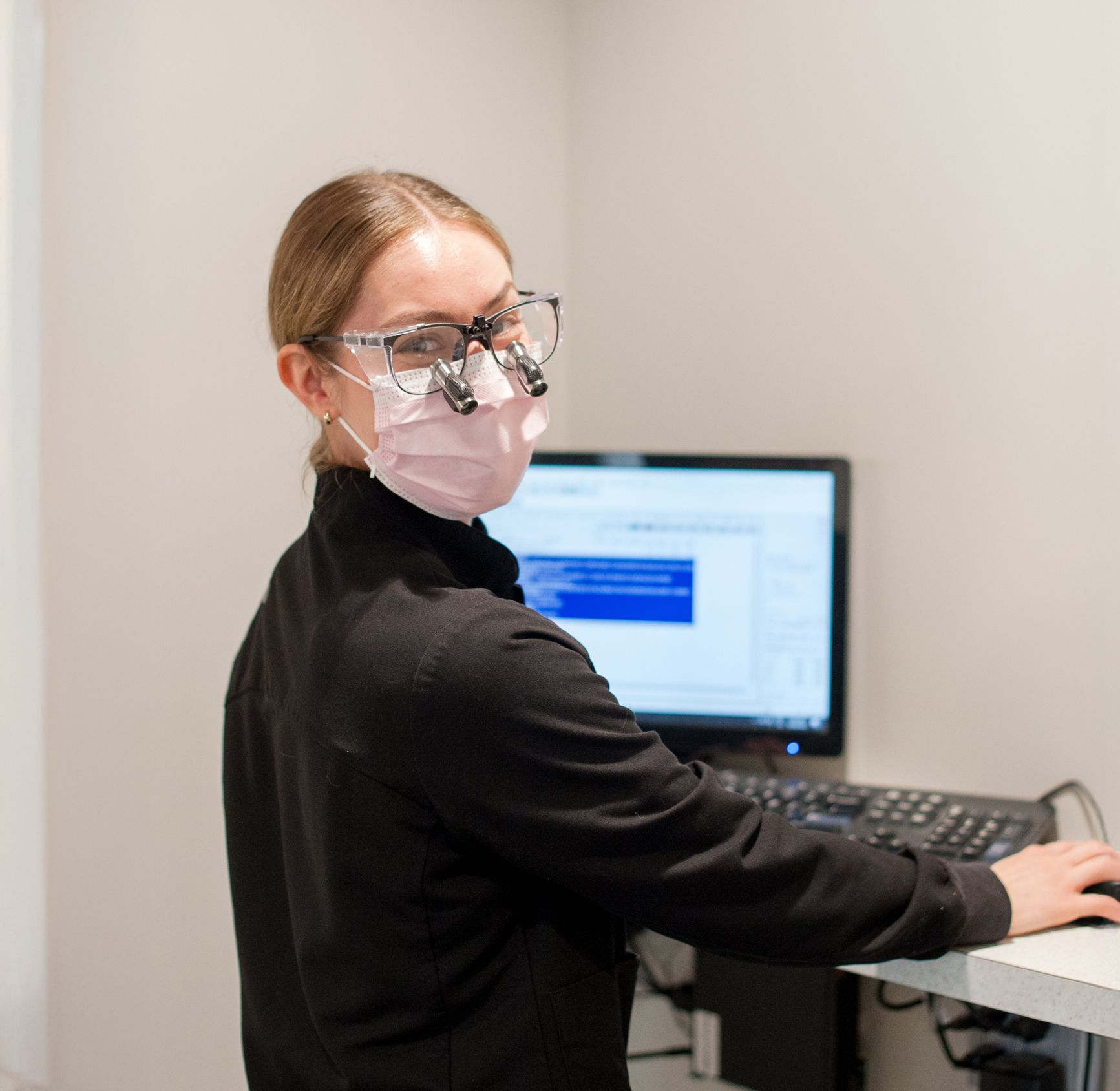 Person in medical scrubs and mask using a computer, wearing surgical loupes.