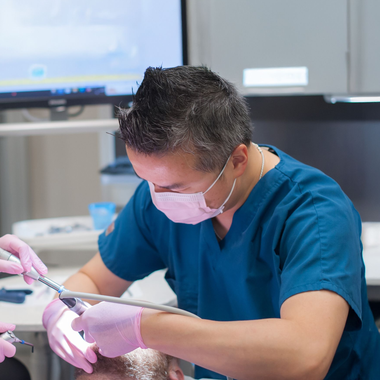 Dentist in burgundy scrubs, mask, and magnification glasses working in a dental office.