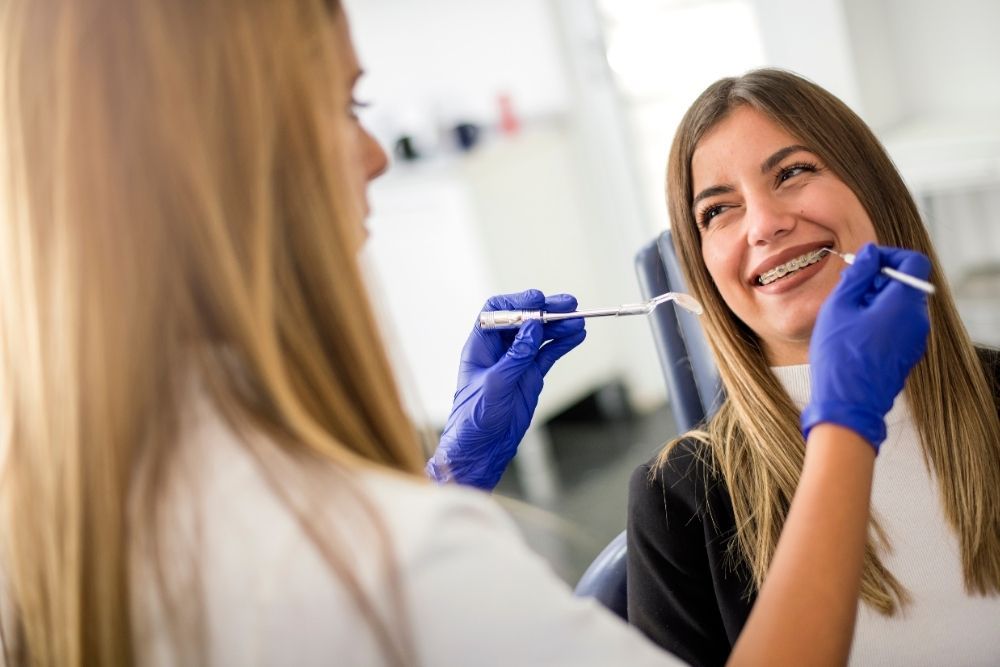Dentist examining patient's teeth with braces, using tools, in an office. Both are smiling.