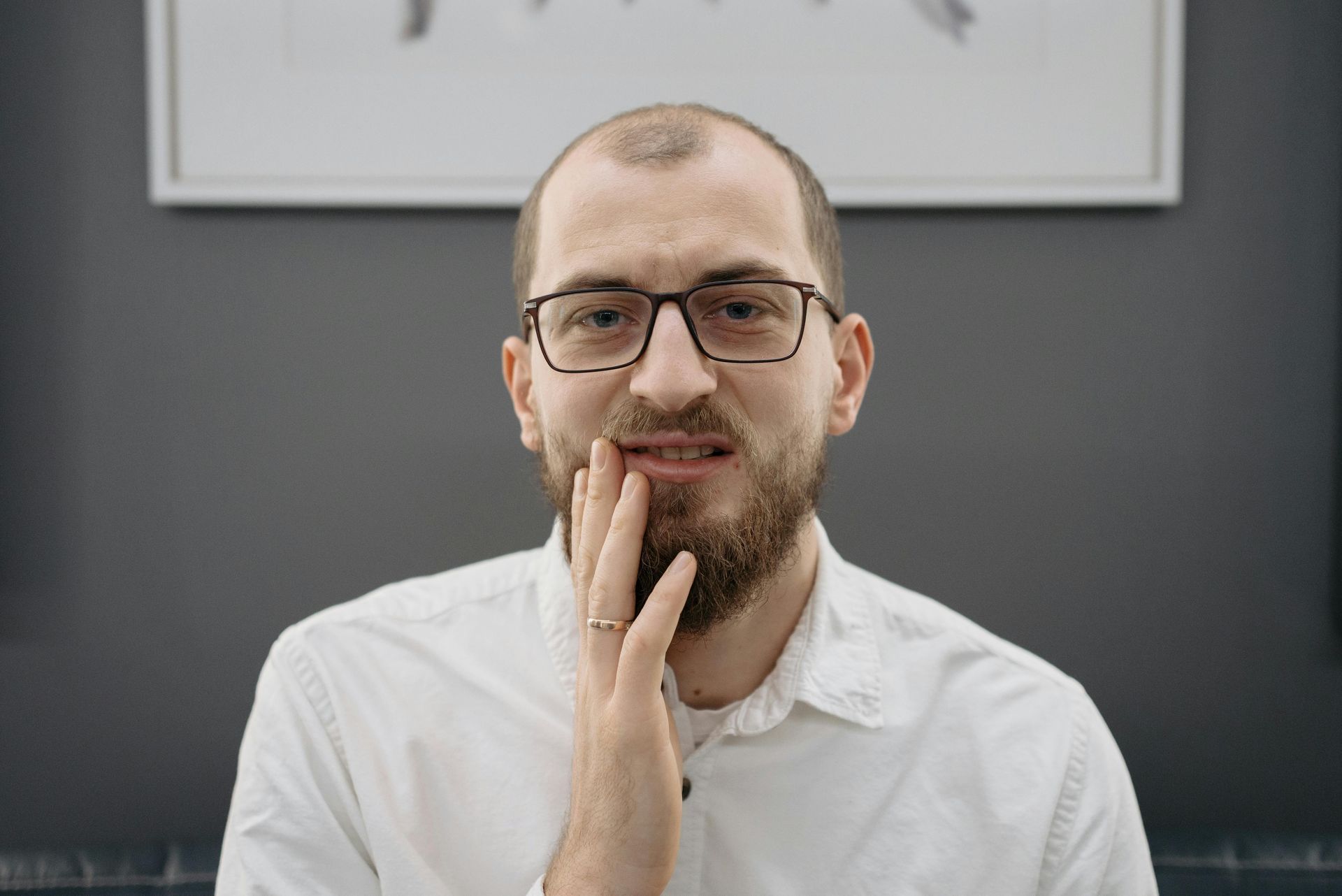 Man in glasses holding his jaw, showing a pained expression. He wears a white shirt against a gray wall.