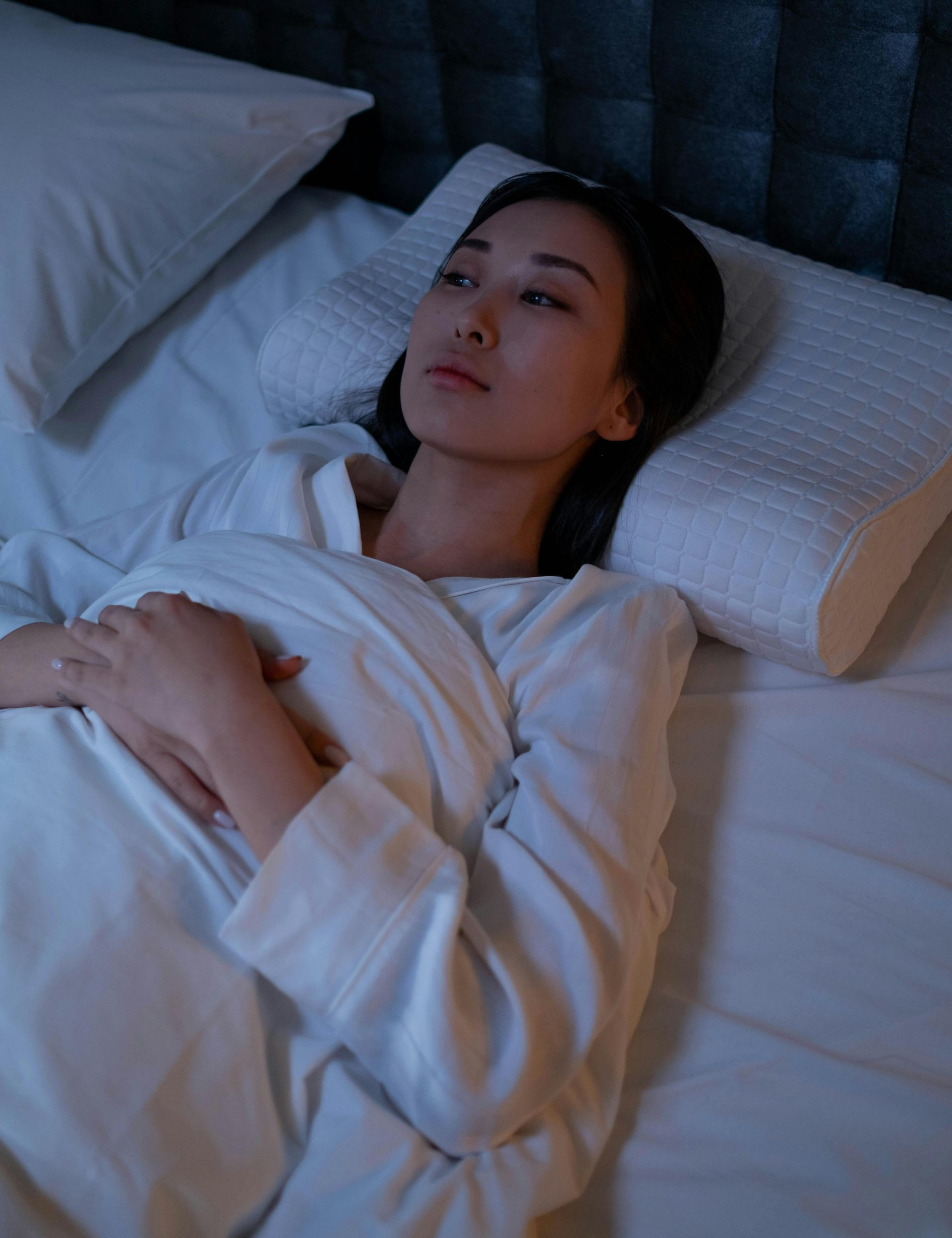 Woman in white robe lies awake in bed, looking thoughtful, illuminated by soft light.