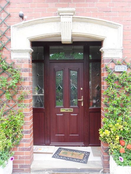 Red front door with glass panels and stone archway, framed by brick walls and greenery.