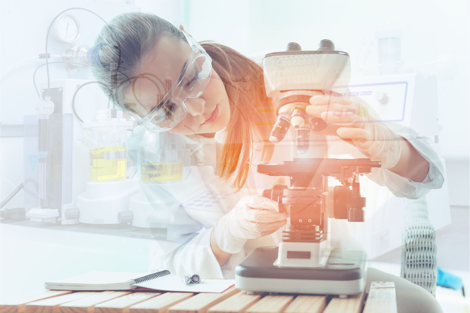 Scientist in lab coat and safety glasses looking through a microscope, light-filled laboratory.