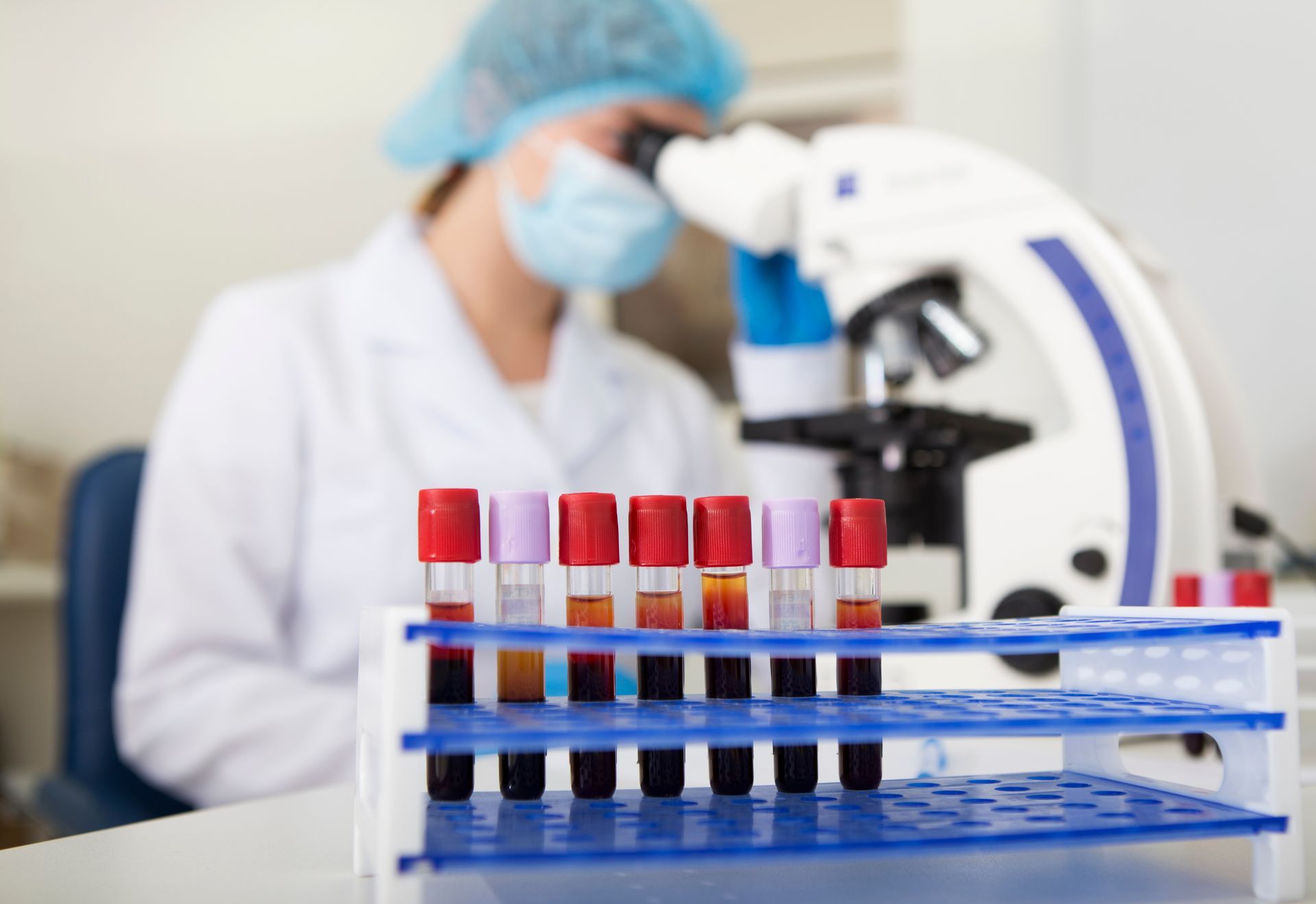 Lab worker in mask looks through microscope, blood sample tubes in rack on table.