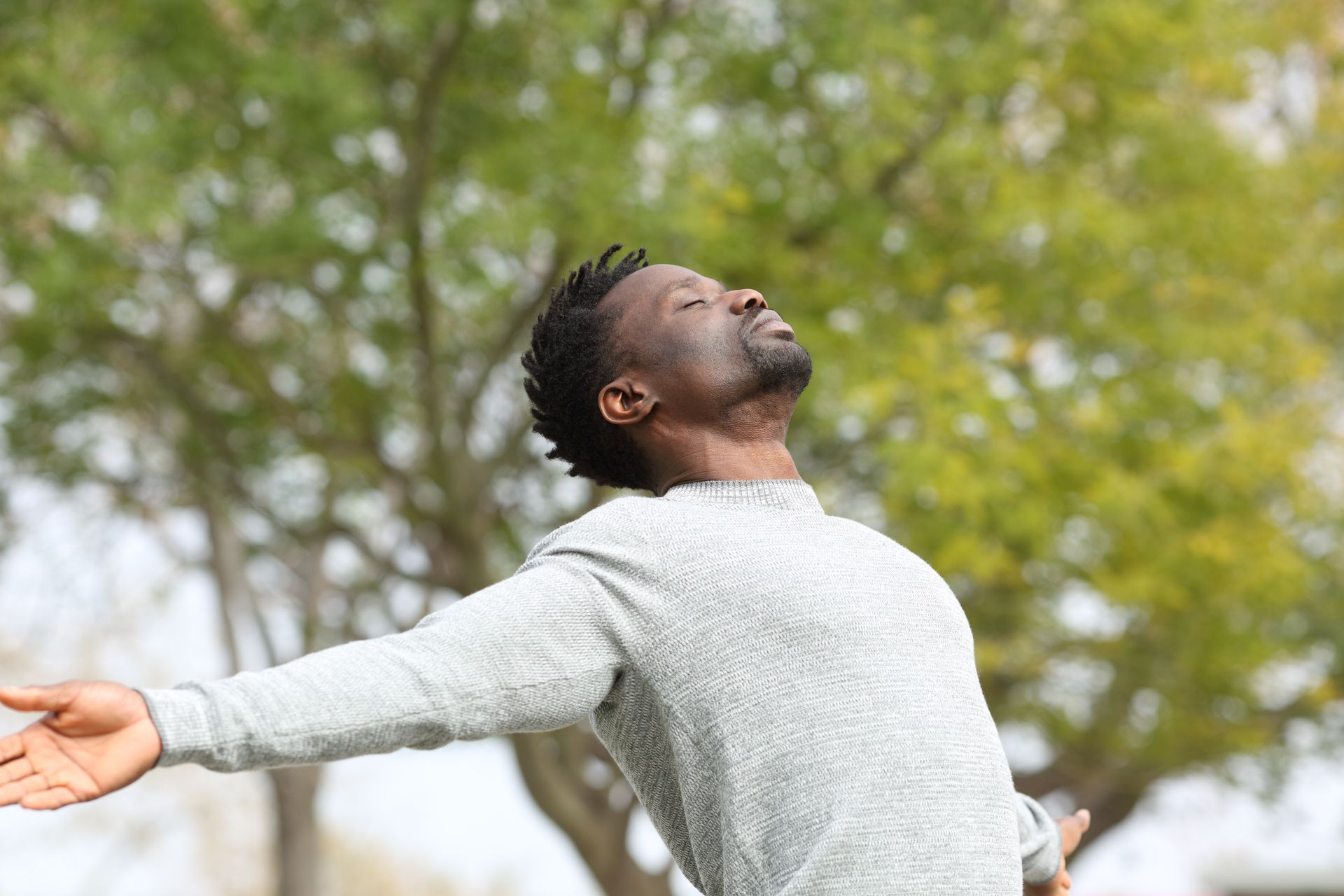 Man with arms outstretched, eyes closed, enjoying the outdoors under a tree, appearing relaxed.