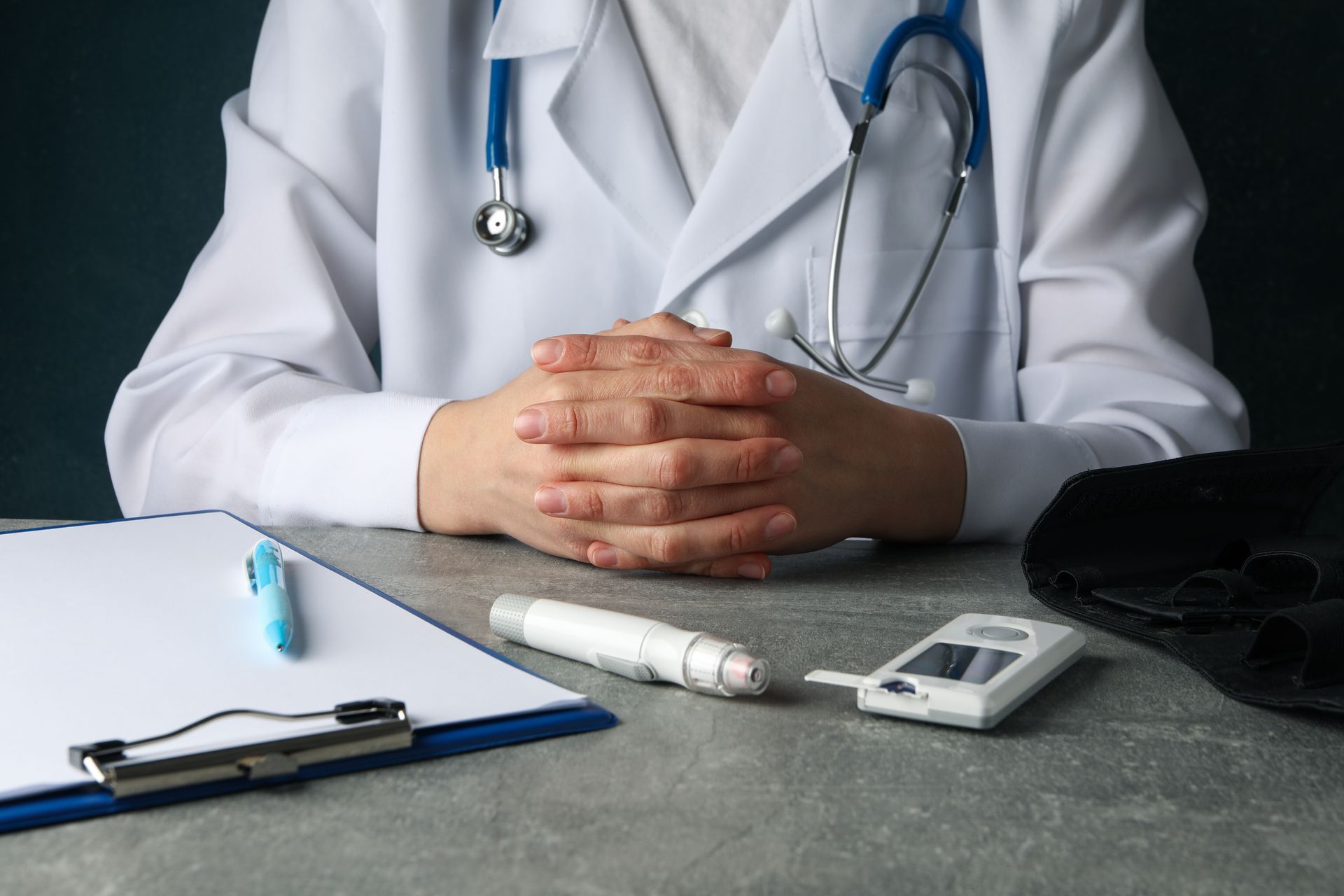 Doctor with stethoscope and clasped hands at desk with medical supplies, clipboard.