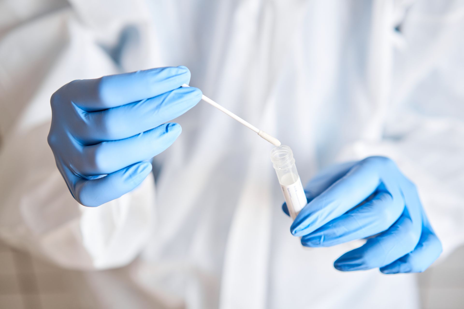 Gloved hands holding a swab and a test tube; person in white lab coat.