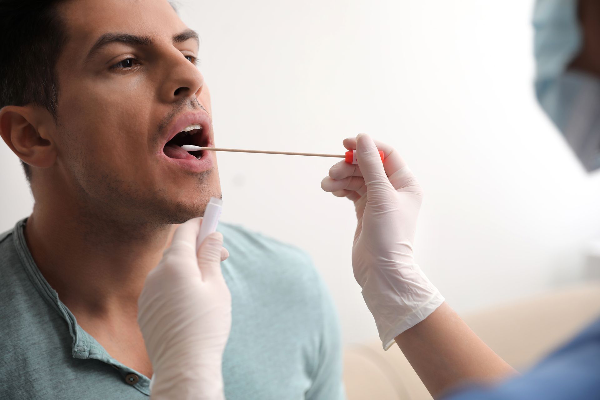 Man receiving throat swab test from medical worker in a white-walled setting.