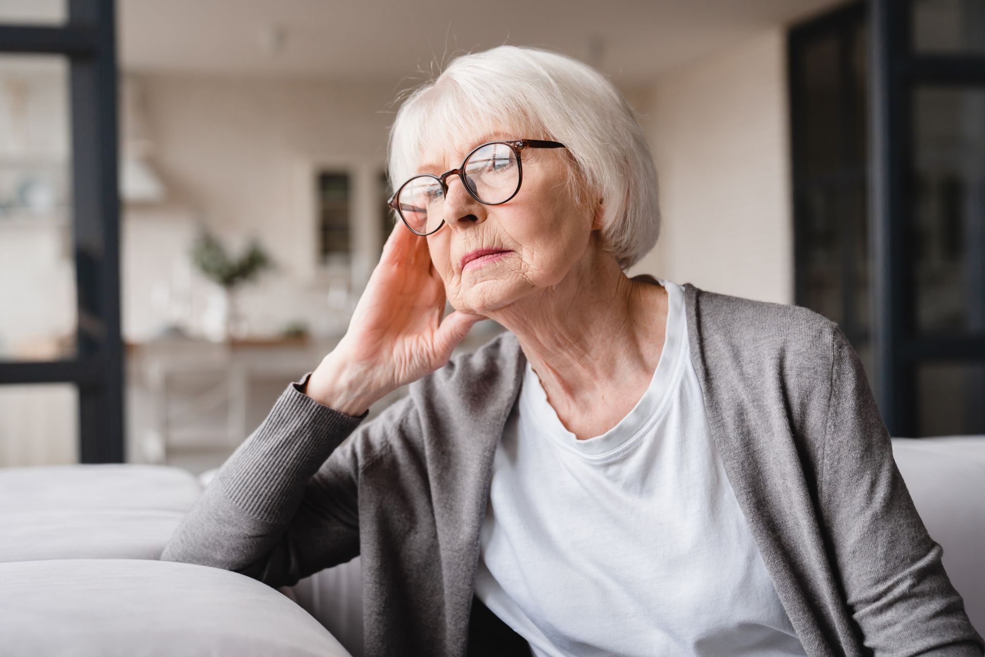 Older woman with glasses, resting head on hand, appears thoughtful, indoors on couch.