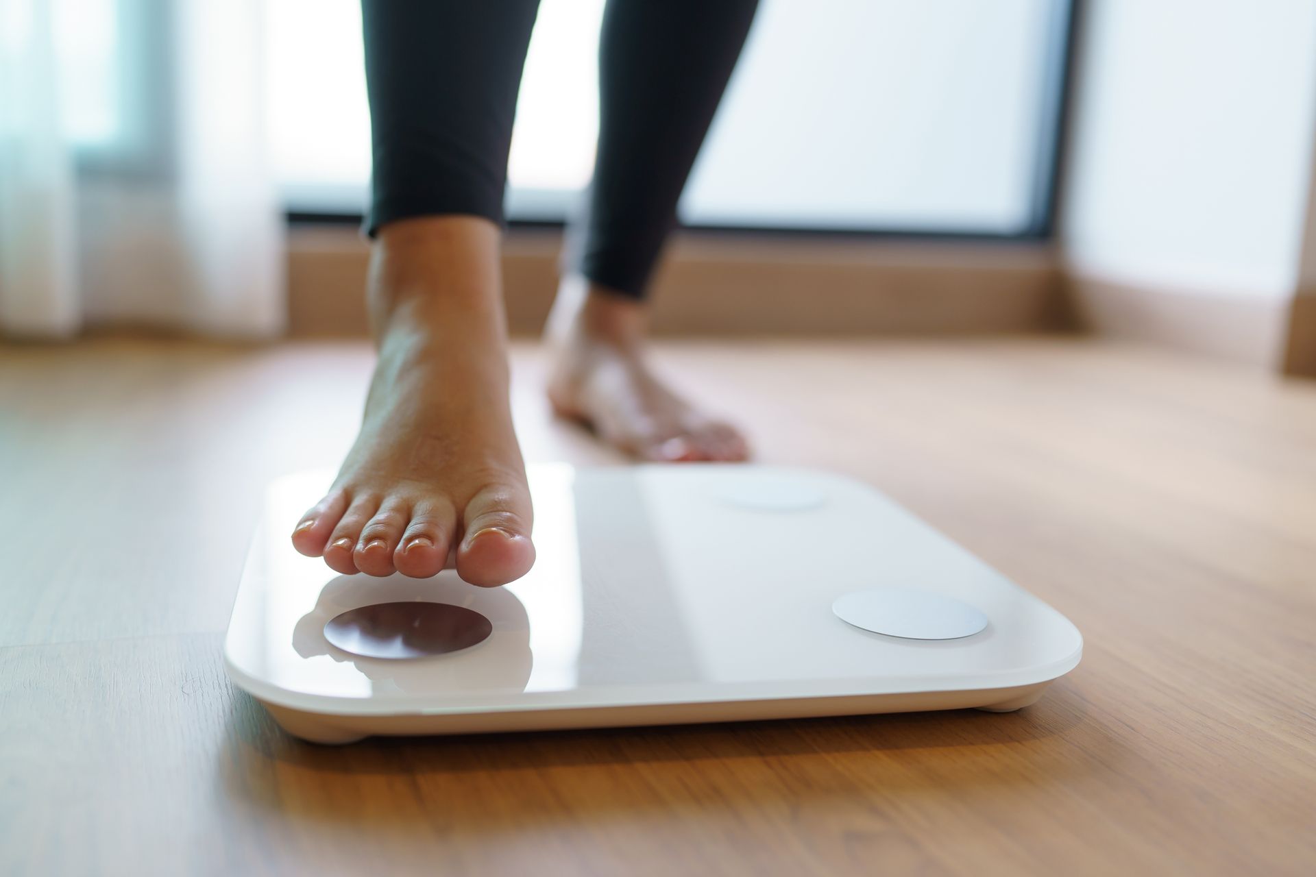 Person's bare foot stepping onto a white scale, indoors, wearing black pants, on a wooden floor.