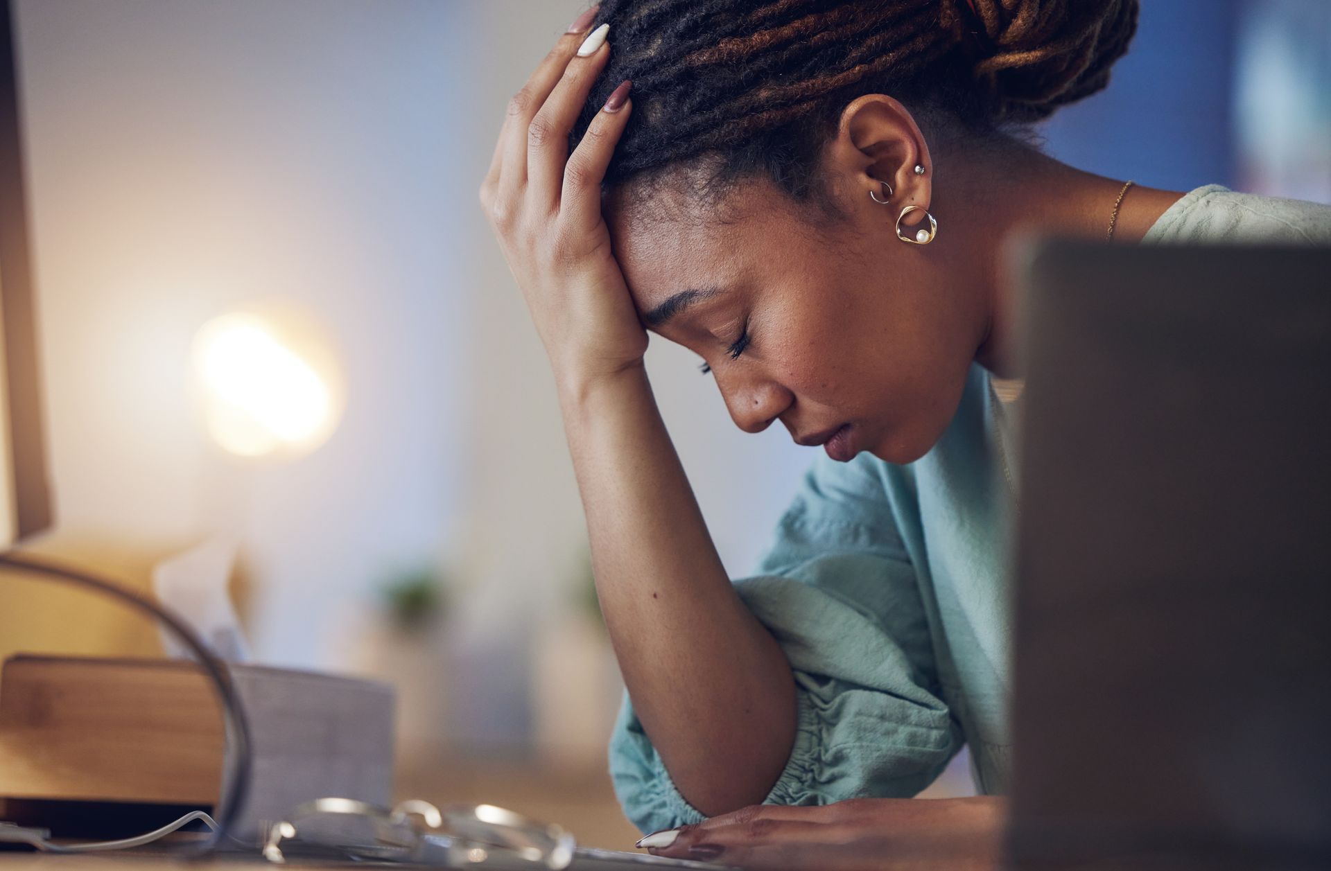 Woman with head in hand, appears stressed at desk, near laptop and lamp.