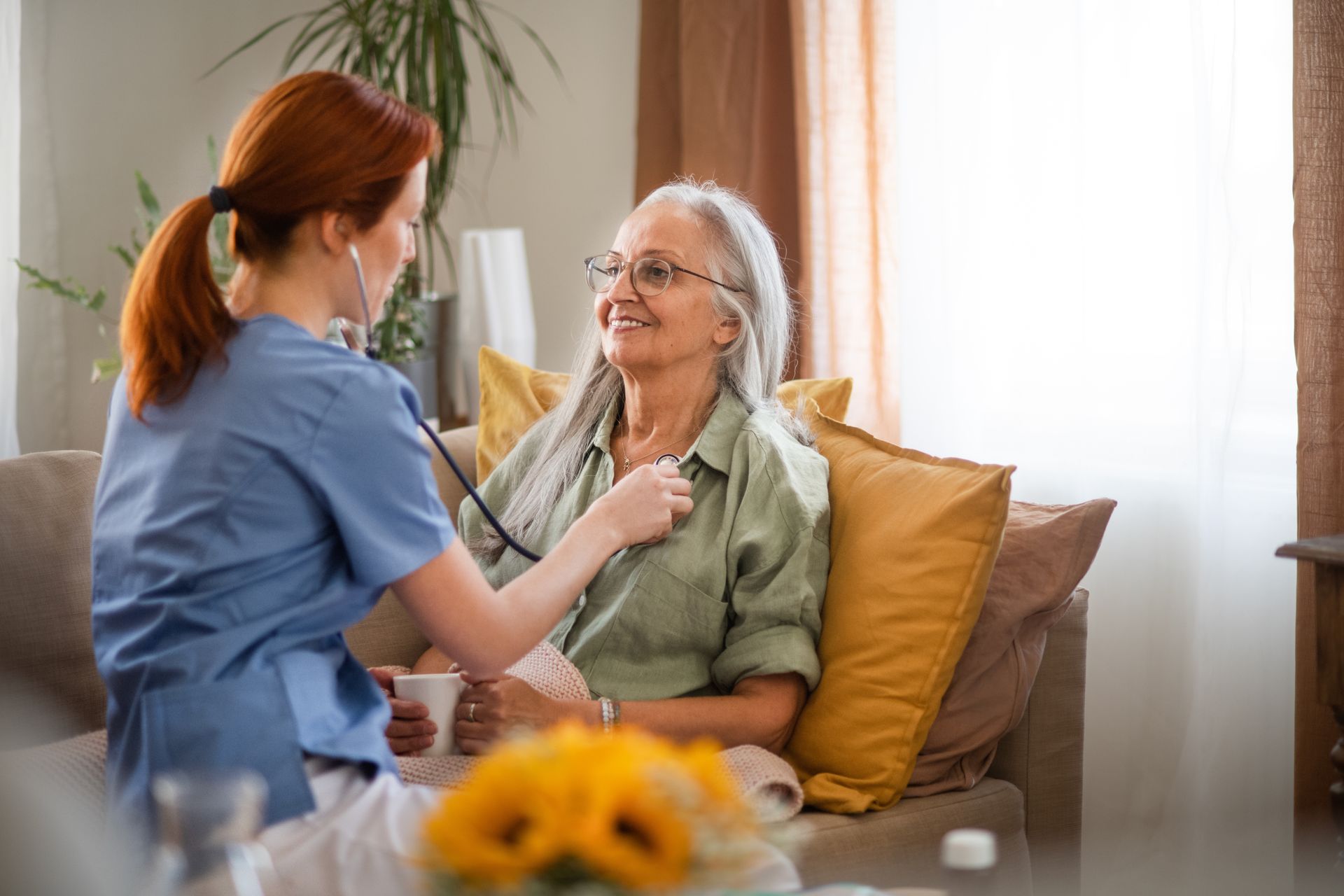 Nurse using stethoscope on elderly patient seated on a sofa.