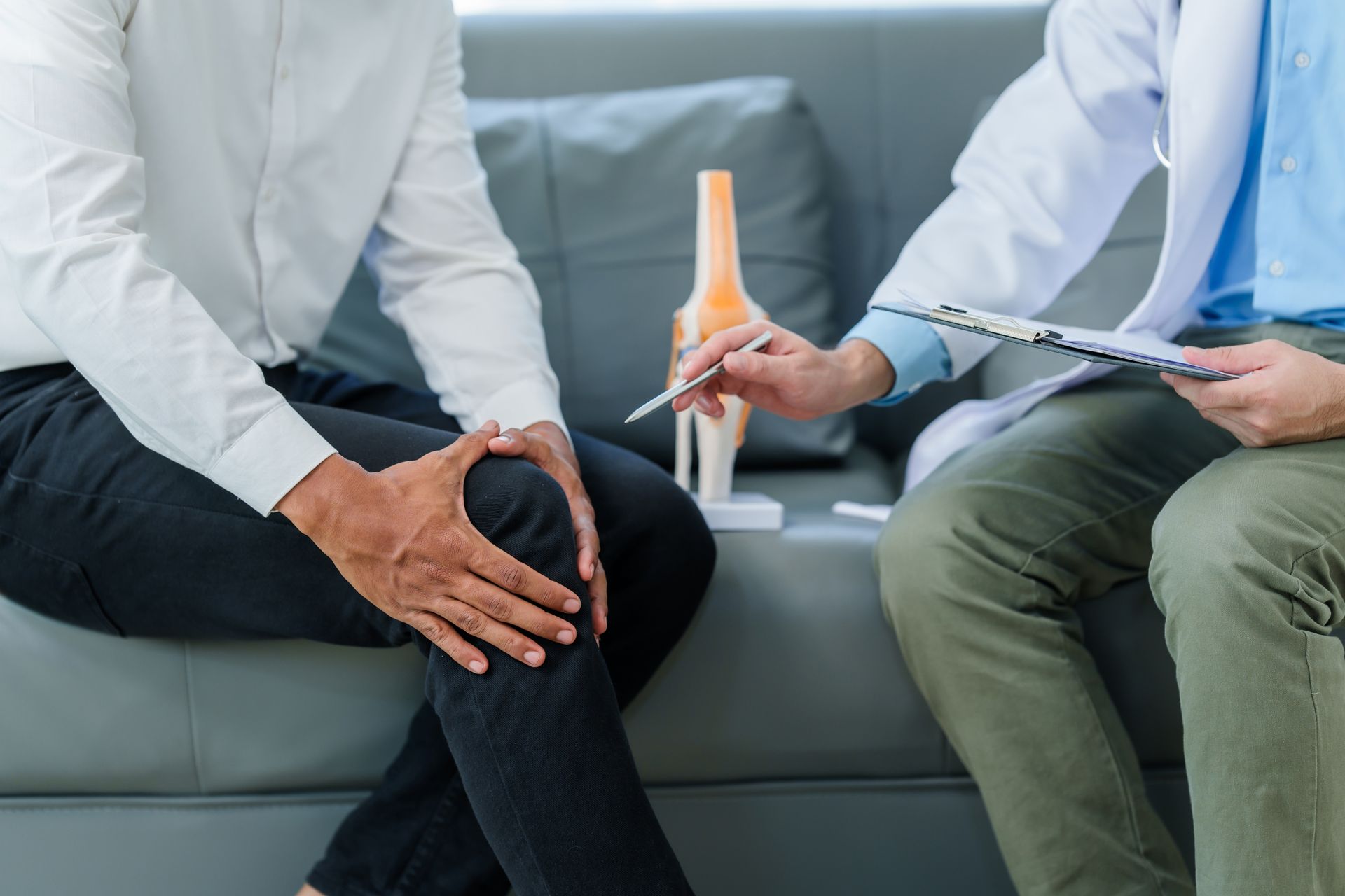 Man points to knee while consulting with doctor, knee model on table.