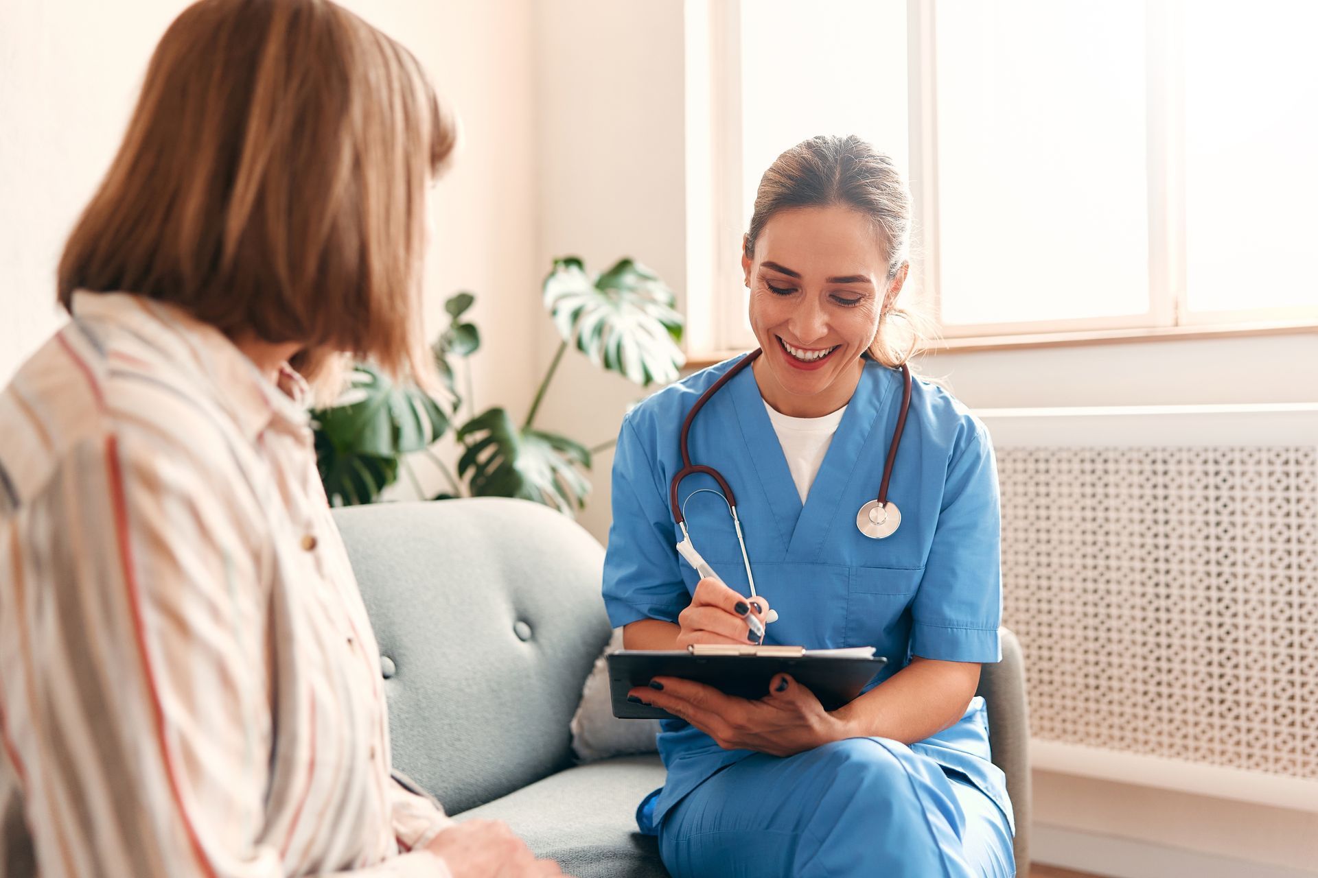 Nurse in blue scrubs, smiling, taking notes while seated and speaking with a patient.