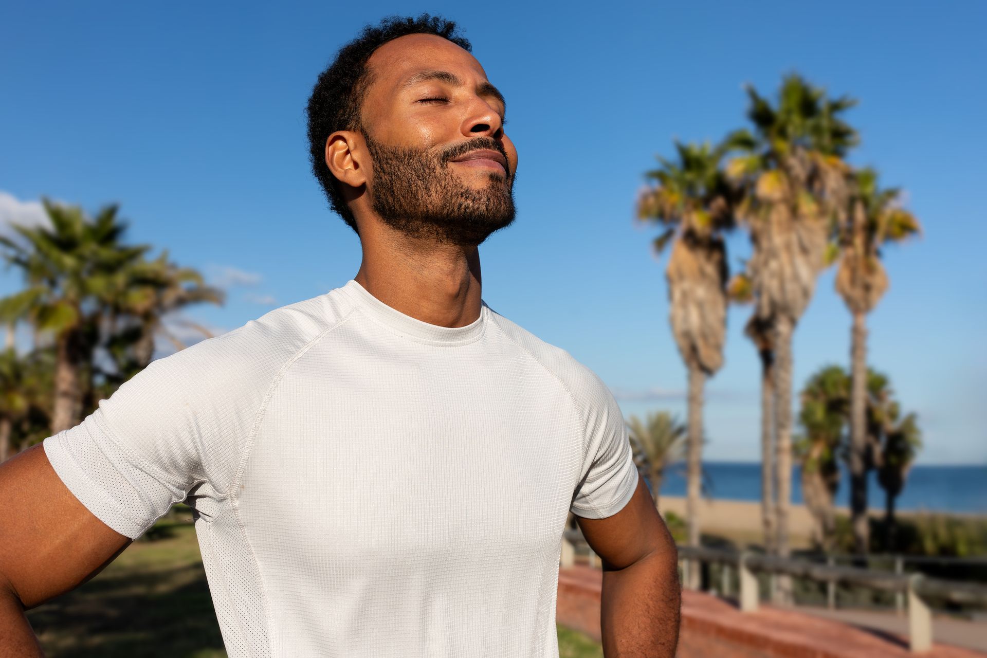 Man with closed eyes, hands on hips, enjoying the sun on a beach with palm trees.