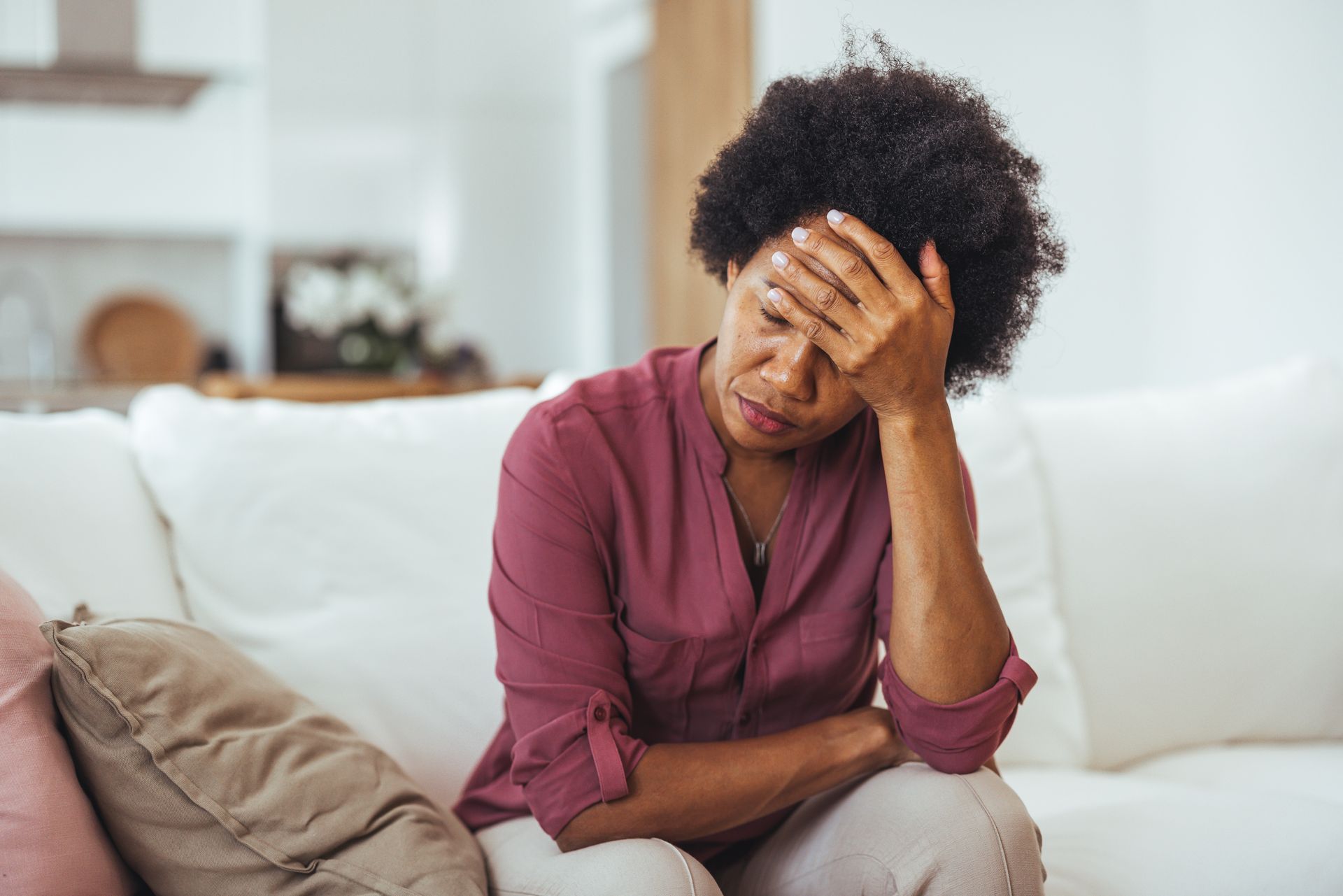 Woman with hand on forehead, looking down, sitting on a white sofa in a light-filled living room.