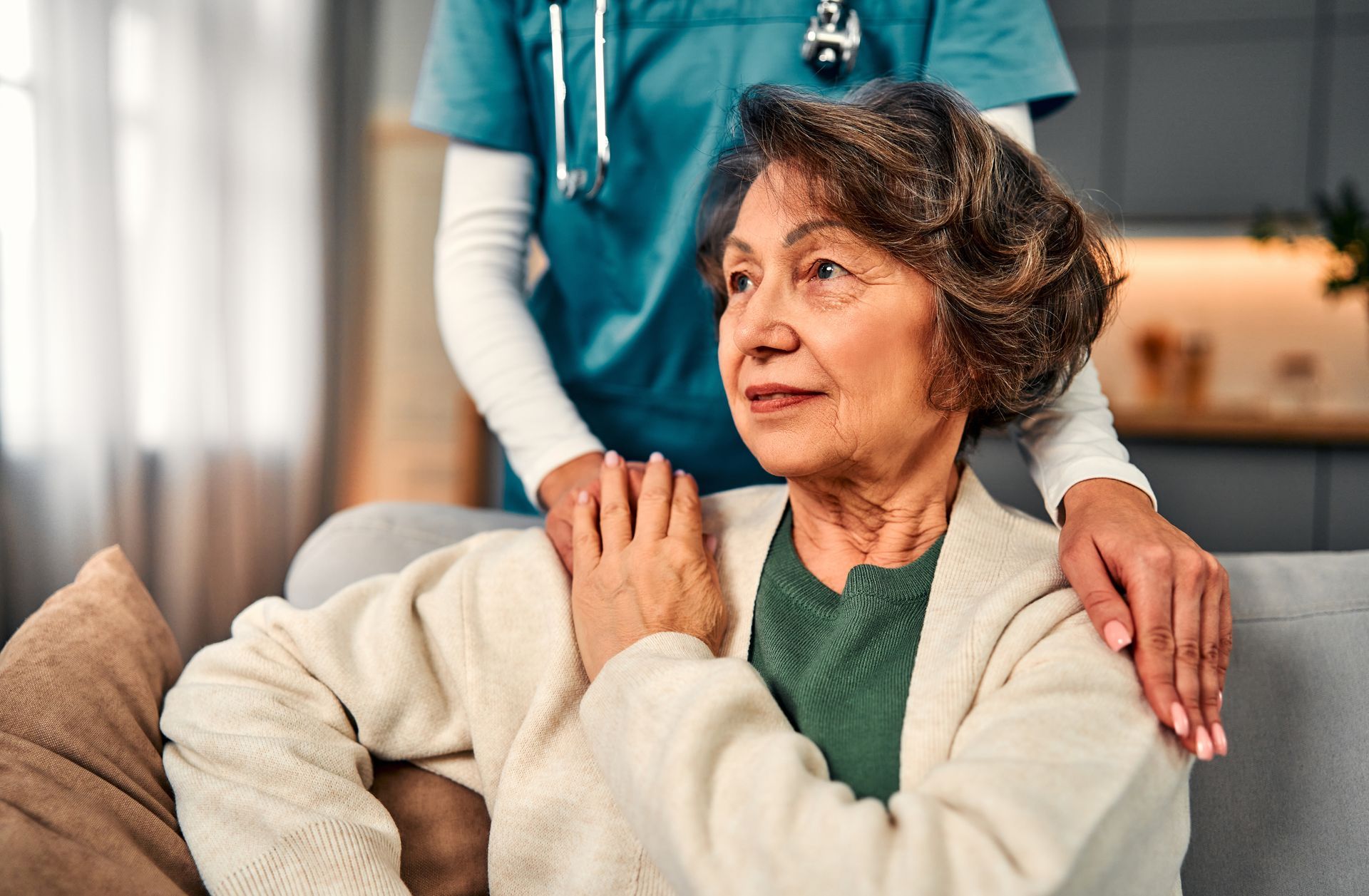 An elderly person sits on a couch, a caregiver's hands on her shoulders. Indoors, natural light.