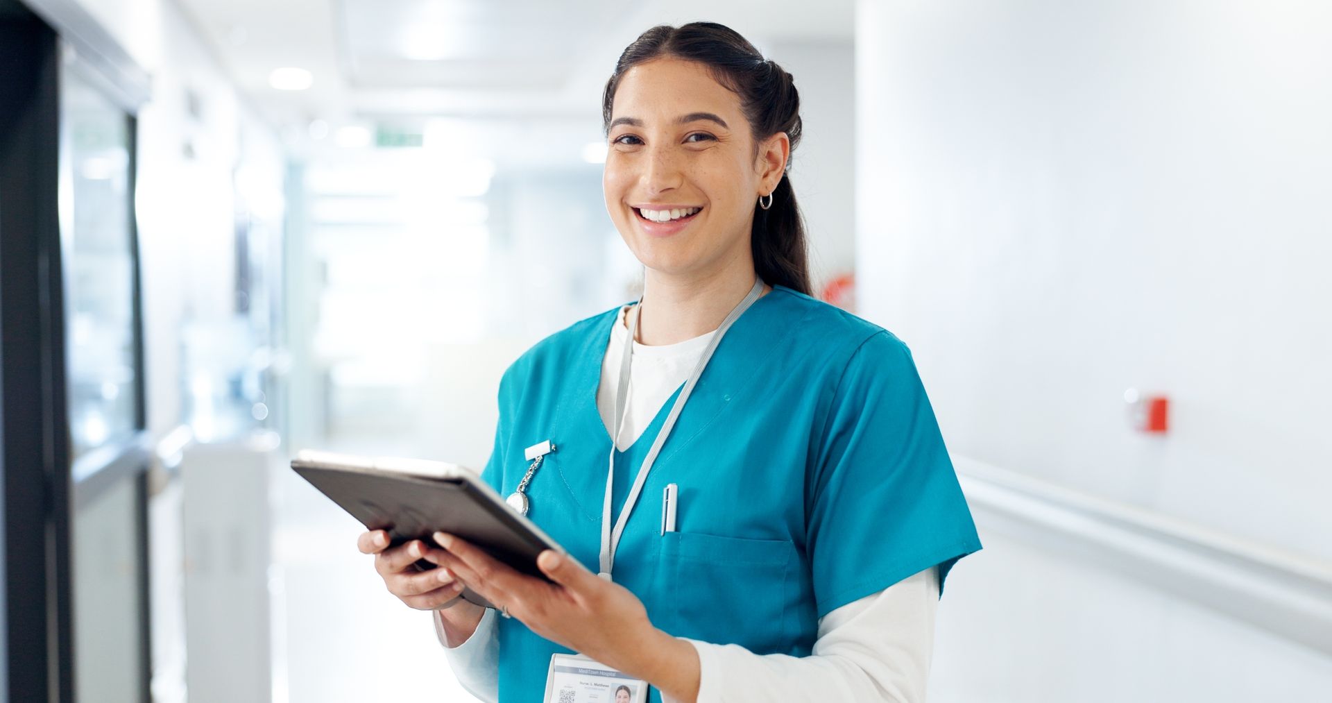 Nurse in blue scrubs smiles, holding tablet, in a hospital hallway.