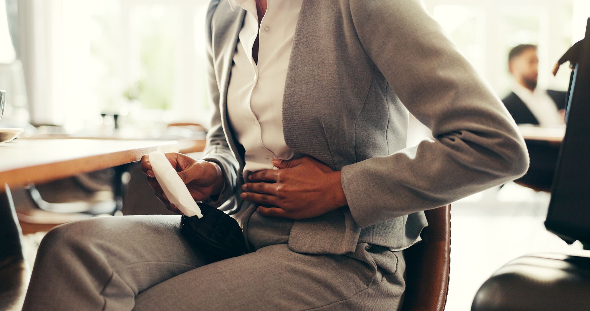 Woman in gray suit, holding stomach, possibly in pain, at a desk.