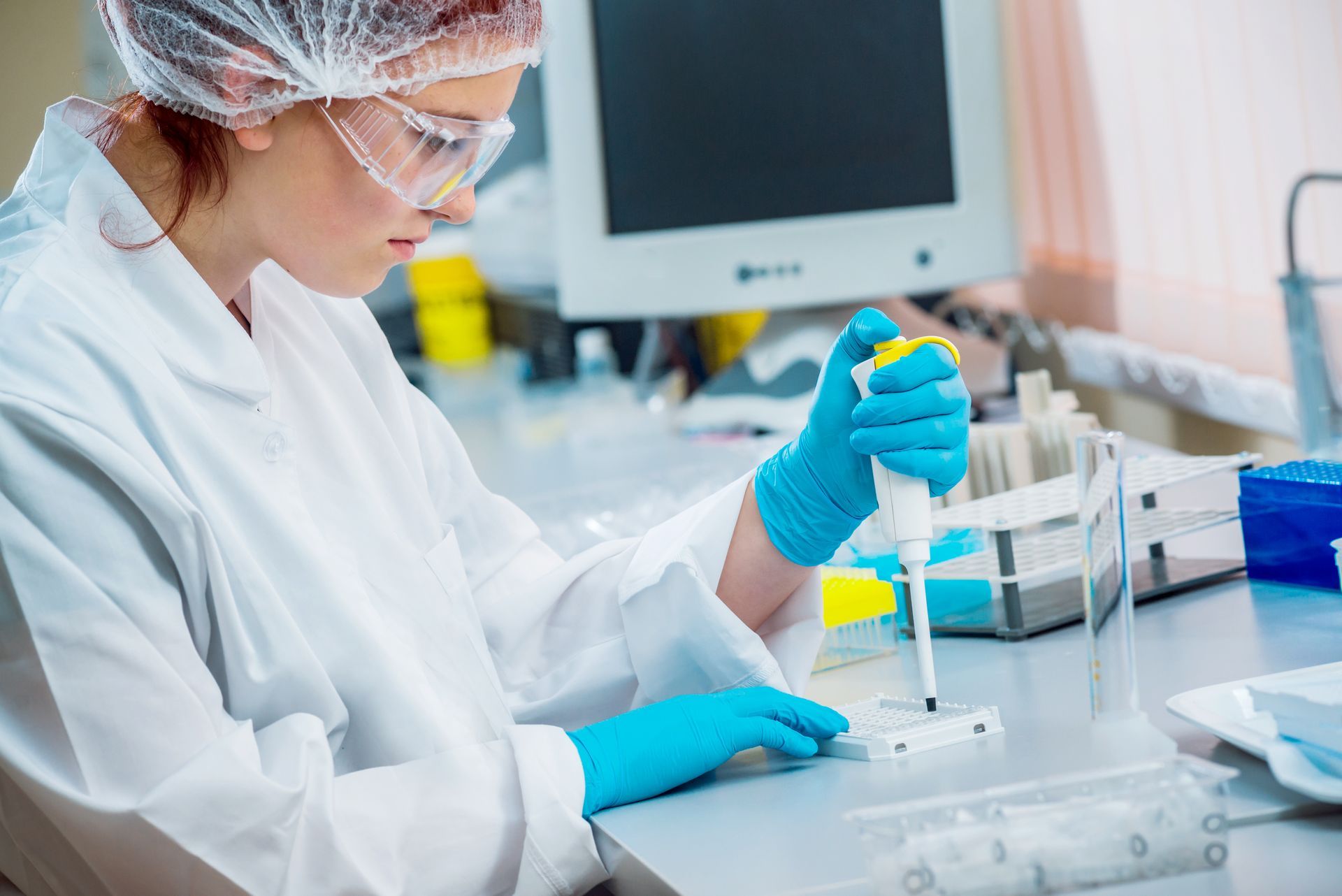 Scientist in lab coat and gloves pipetting liquid into a well plate.