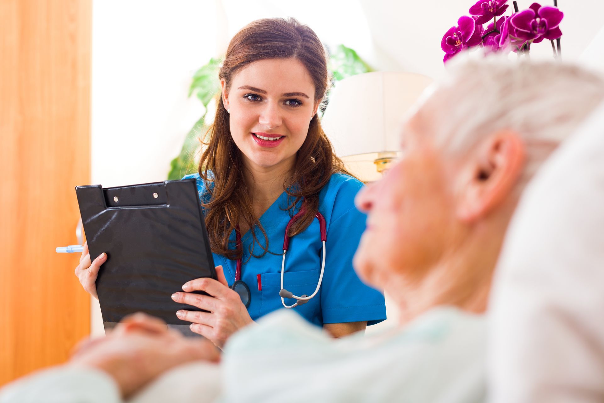 Nurse in blue scrubs with stethoscope, holding a clipboard, speaking to a patient in bed.