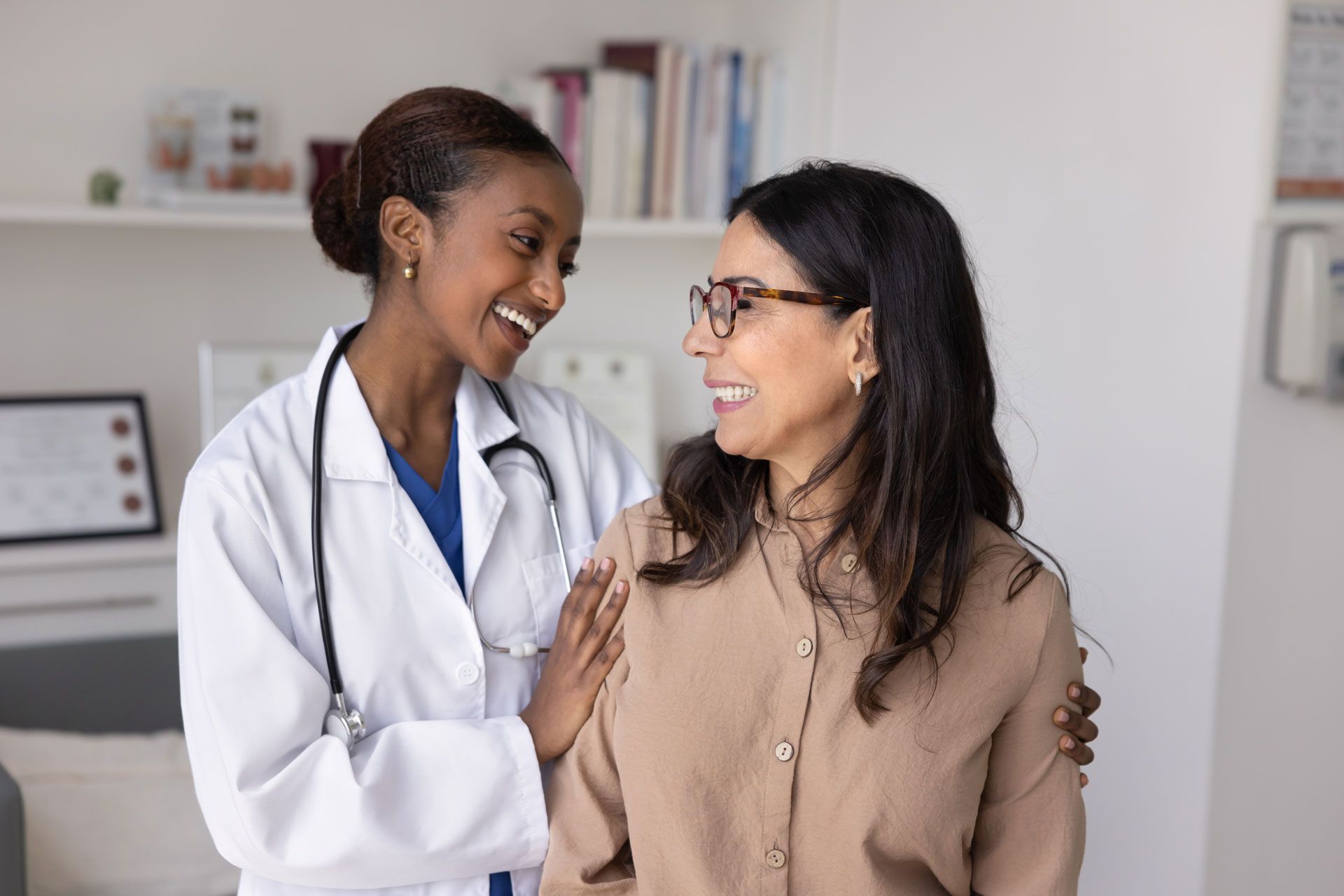 A smiling healthcare professional in a lab coat with a stethoscope places a hand on the shoulder of a smiling patient.