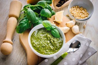 Una ciotola di pesto verde con basilico, aglio, pinoli, formaggio e un pestello di legno su un tavolo da cucina.