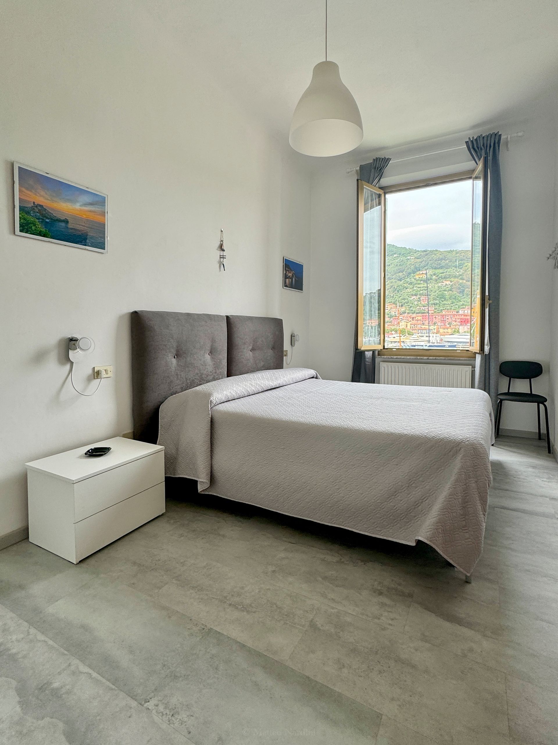 Camera da letto luminosa con letto grigio, pareti bianche, ampia finestra e vista sulla città .
