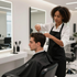 A hairstylist in a black apron trims the hair of a person sitting in a salon chair in front of a mirror.
