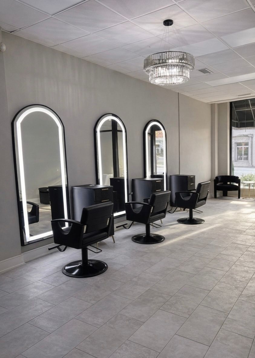 A hair salon featuring three stations with illuminated arched mirrors, black chairs, and a contemporary crystal chandelier.