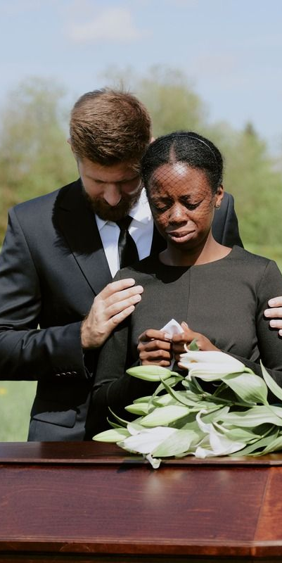 Mourning woman crying, comforted by a person in a suit, at a funeral. Flowers rest on a casket.