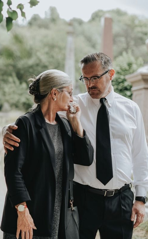 A woman cries as a man comforts her at a cemetery. Both are dressed in black.