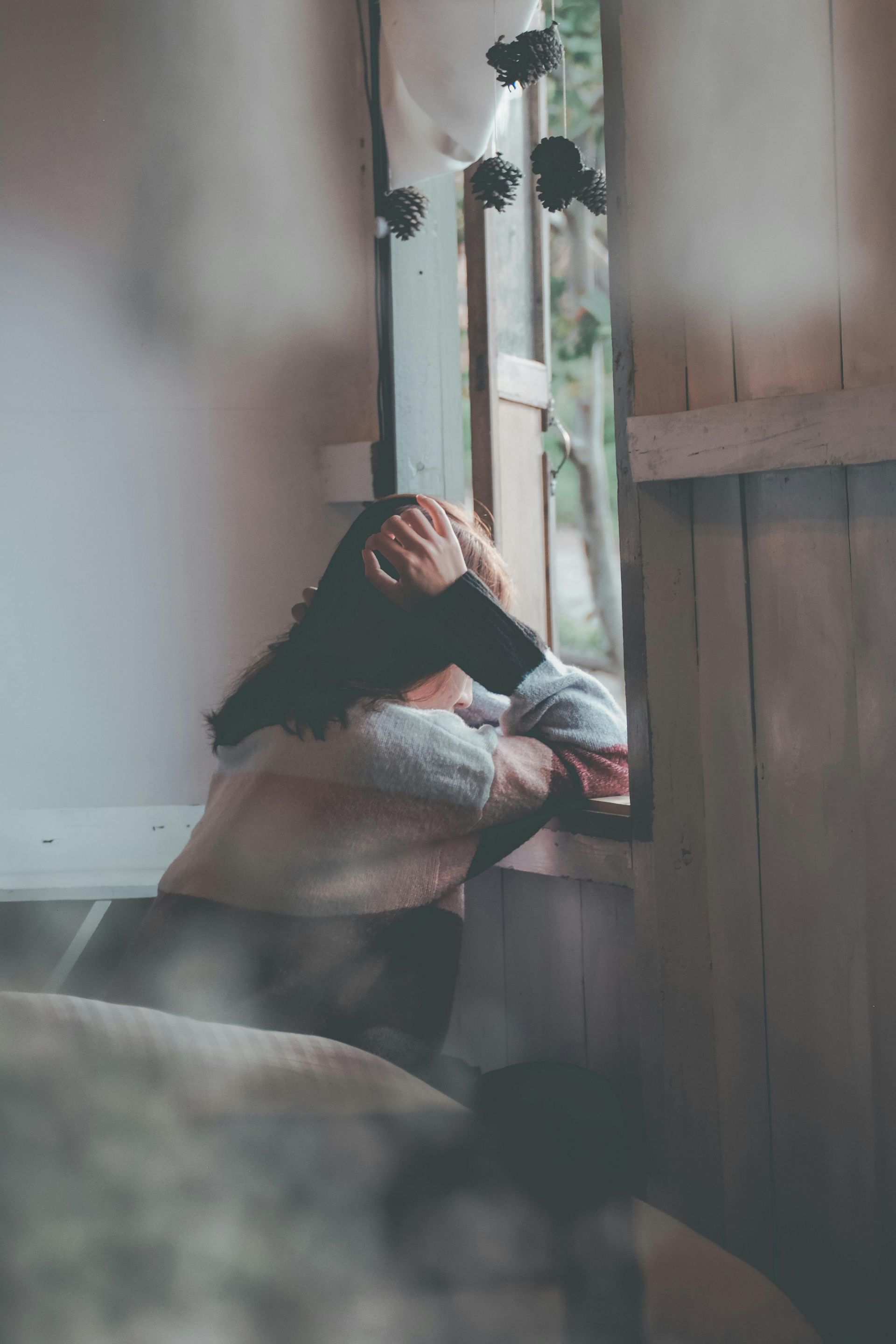 Person with dark hair, head in hands, looking out a window. Interior shot with wooden framing.