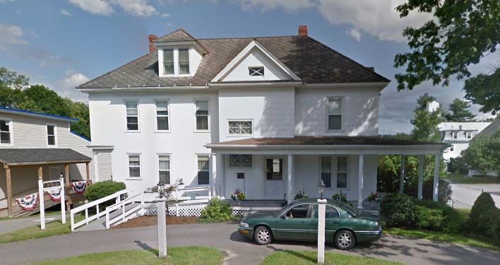 White two-story house with porch; green car in driveway; ramp leading to entrance; sunny day.