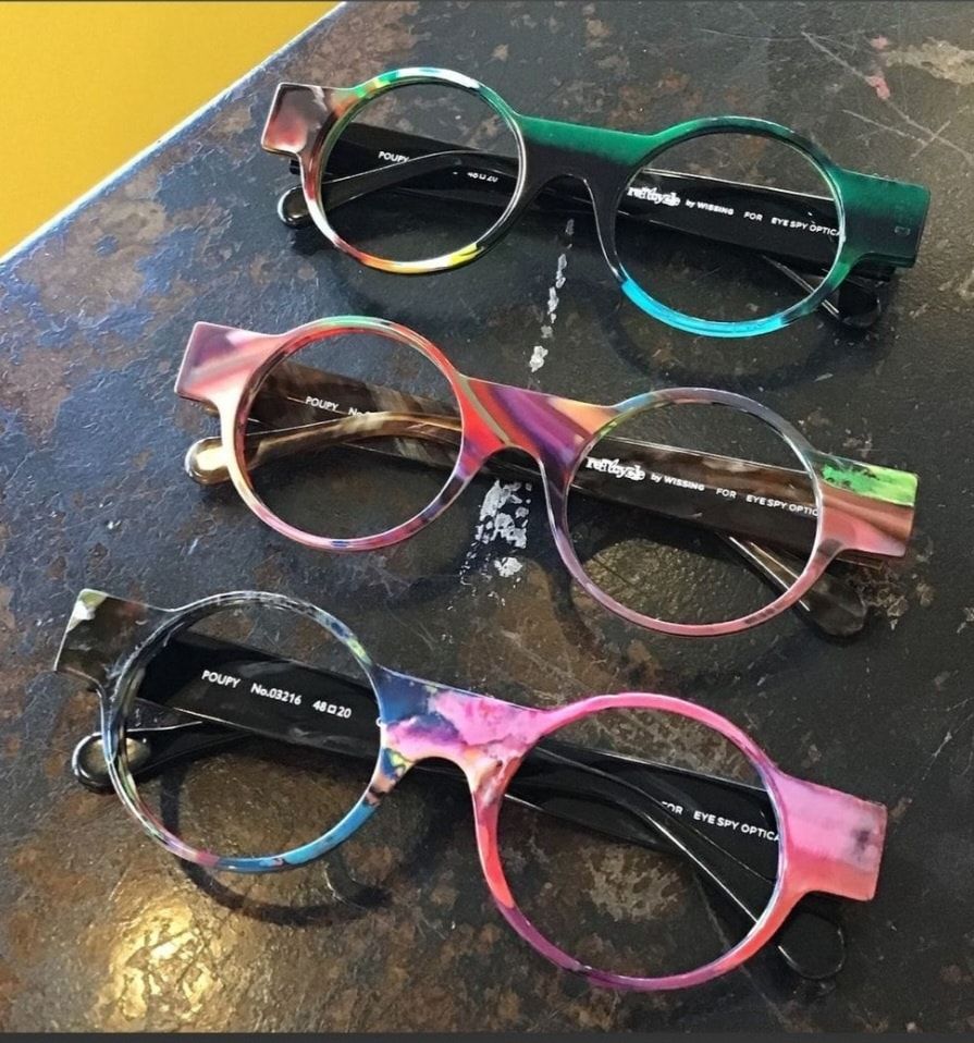 Three colorful eyeglasses frames with round lenses, stacked on a textured, dark surface.