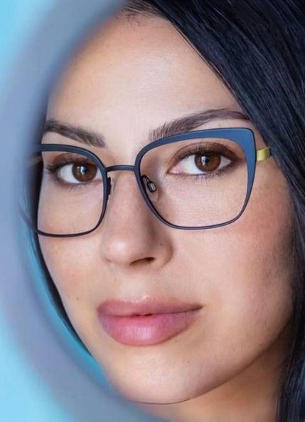 Woman wearing glasses, looking through a light blue frame.  She has dark hair and a neutral expression.