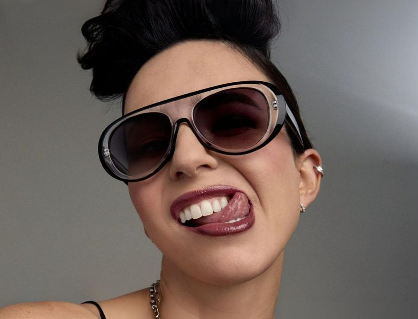 Katy Perry with sunglasses, sticking out her tongue, smiling. Dark hair, lipgloss.