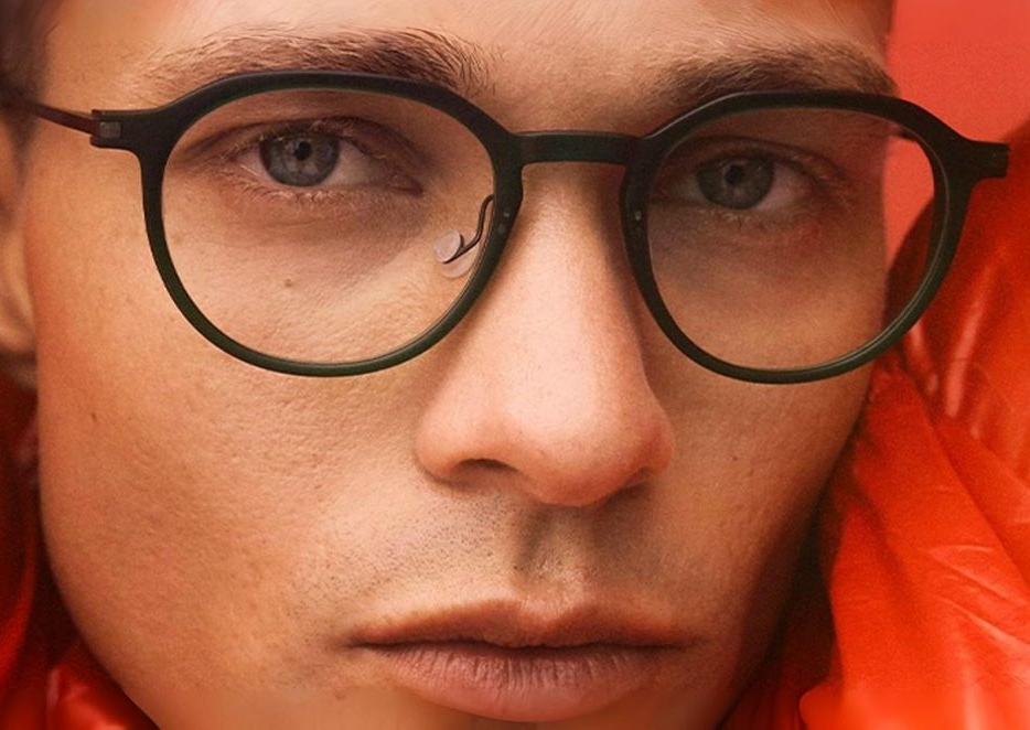 A man wearing dark-rimmed glasses and an orange jacket. His face is centered, with a neutral expression.