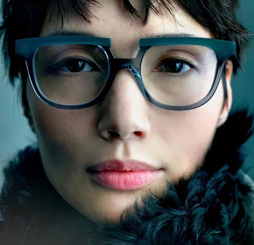 A person wearing glasses, black scarf in a studio setting with a logo in the bottom left corner.