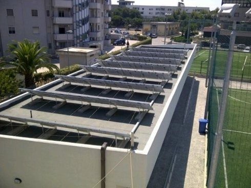 Tetto con pannelli solari; edificio, palma e campo verde sullo sfondo. Giornata luminosa e soleggiata.