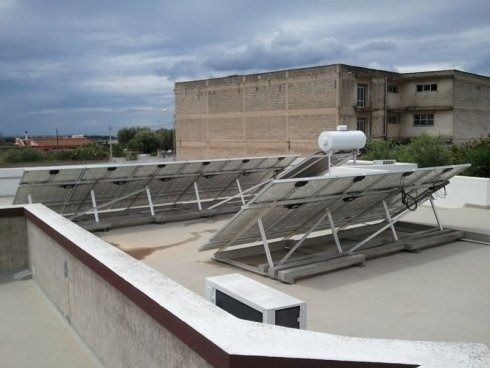 Pannelli solari montati su un tetto piano con un serbatoio d'acqua, sullo sfondo di un cielo nuvoloso e di un edificio beige.