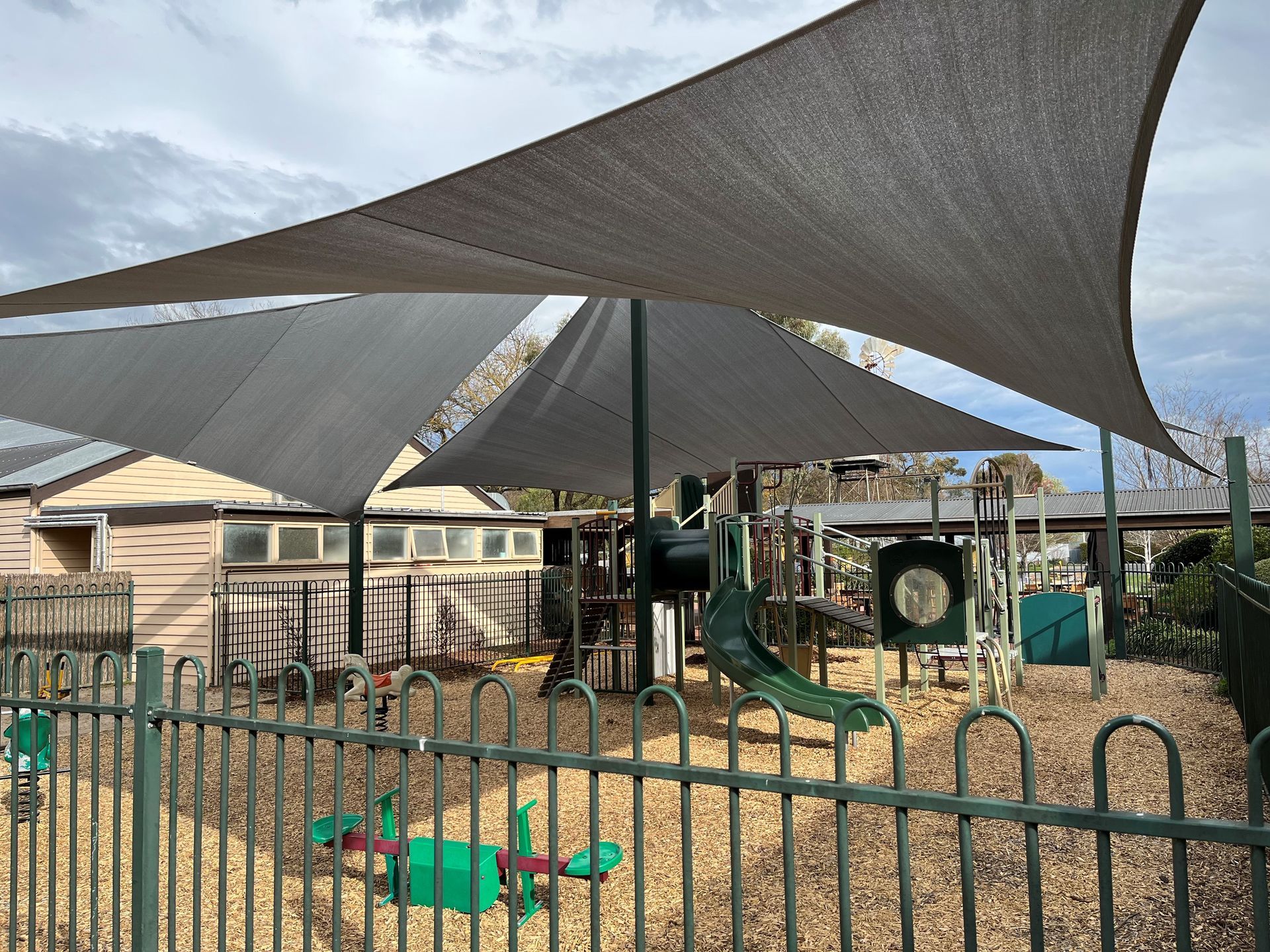 Shaded playground with green climbing equipment and a black fence in front of buildings — The Gazebo and Shade Centre in Jindera, NSW