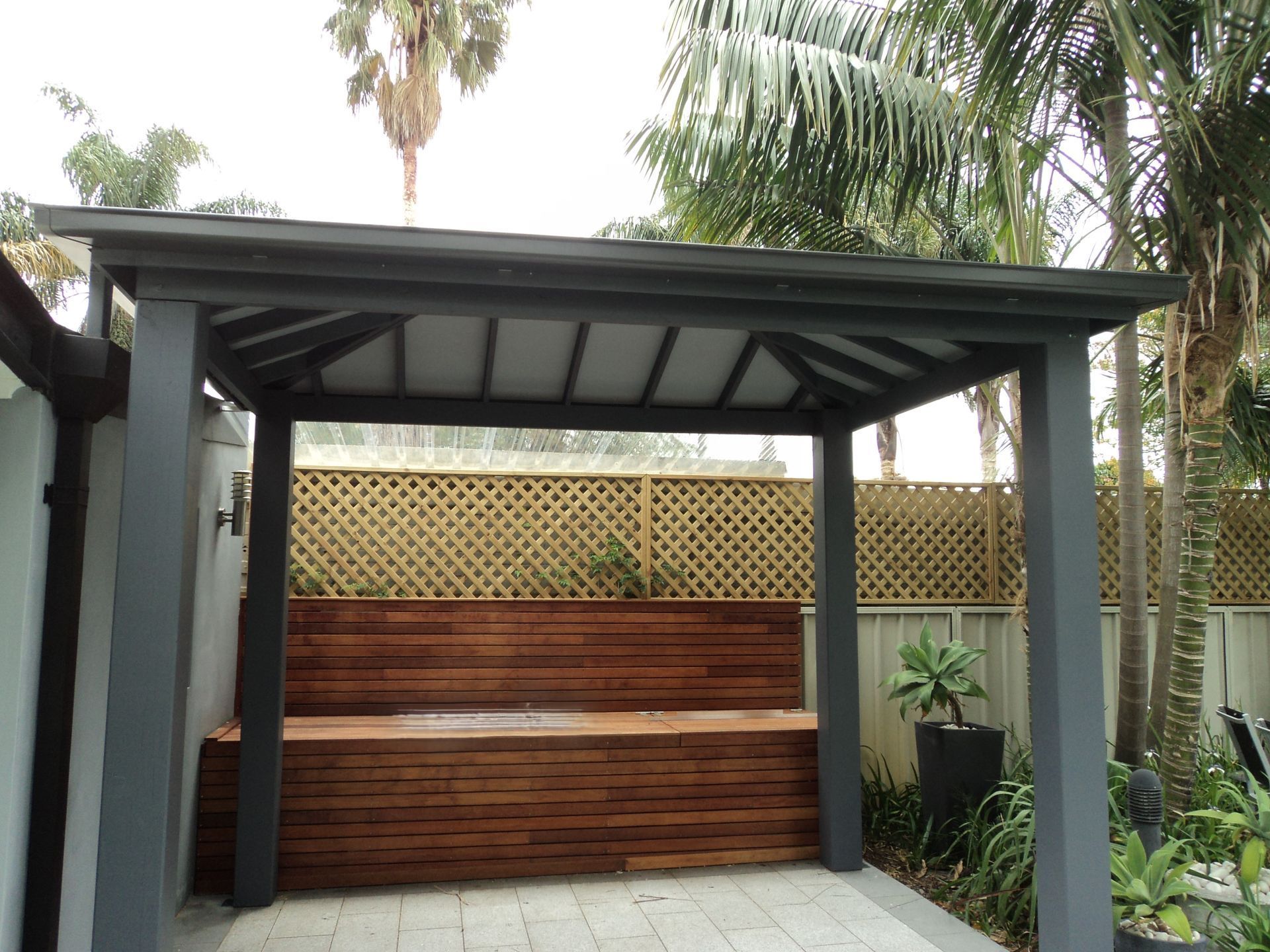 Covered outdoor patio with wood bar counter, lattice fence, and tropical plants under palm trees  — The Gazebo and Shade Centre in Jindera, NSW
