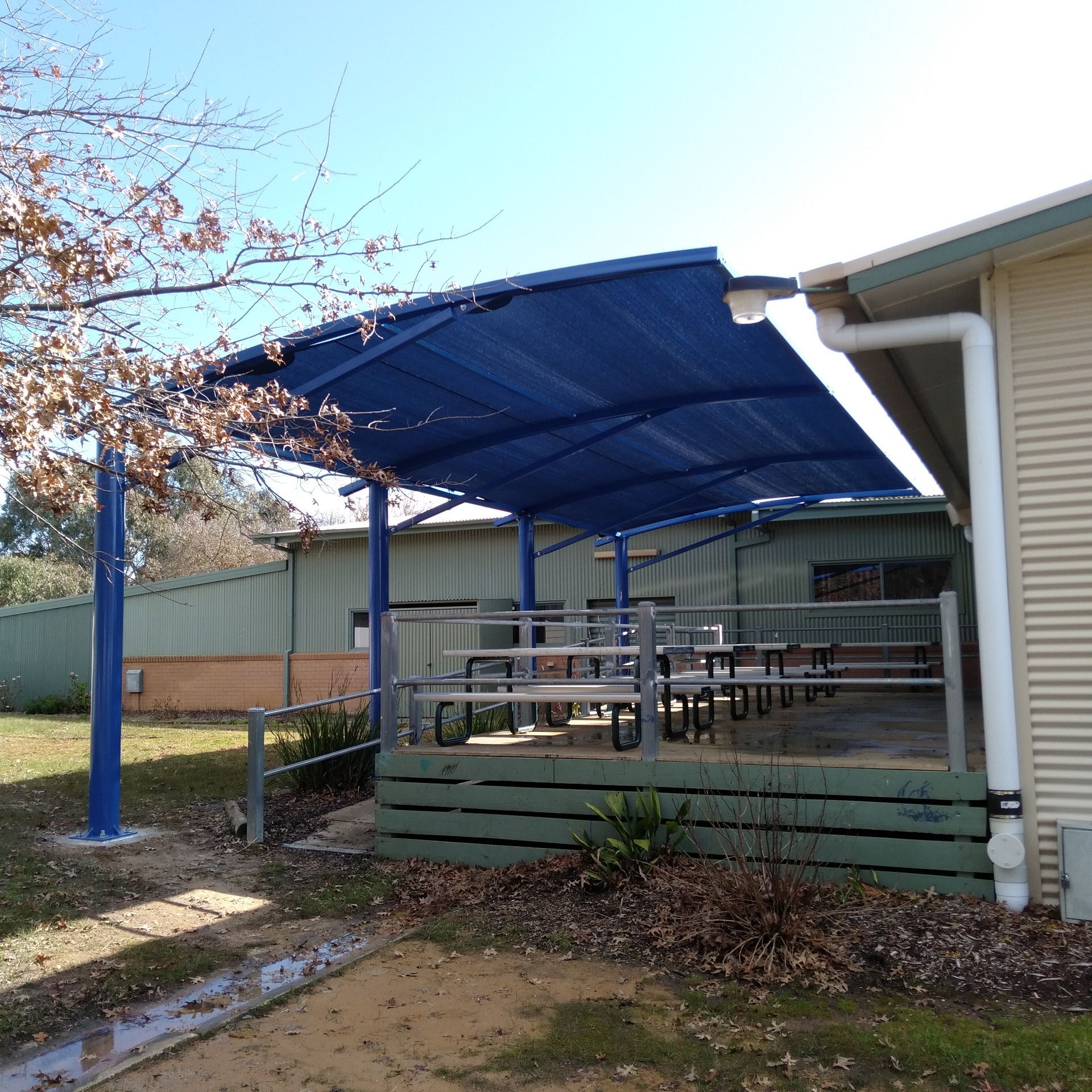 Covered outdoor patio with blue awning, picnic tables, and green skirted deck beside a building — The Gazebo and Shade Centre in Jindera, NSW