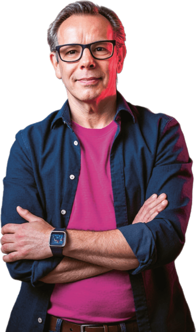 Een persoon met een bril, een roze shirt en een blauw overhemd staat met de armen over elkaar en kijkt naar de camera.