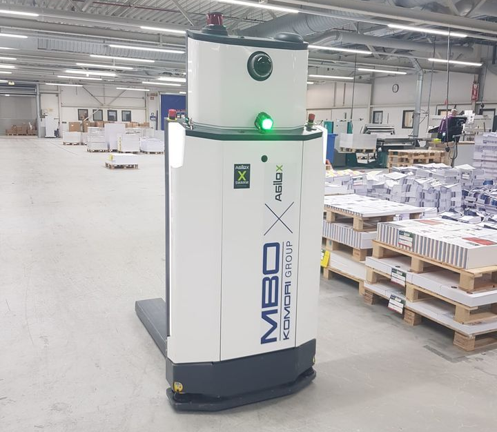 Bureau met laptop, koffie, muis, notitieboekje, pen en handen die een boekje vasthouden, tegen een felgele achtergrond.