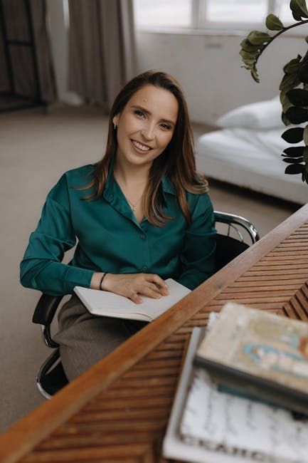 Vrouw in een turquoise blouse glimlacht aan een tafel met een open notitieboekje in een lichte kantooromgeving.