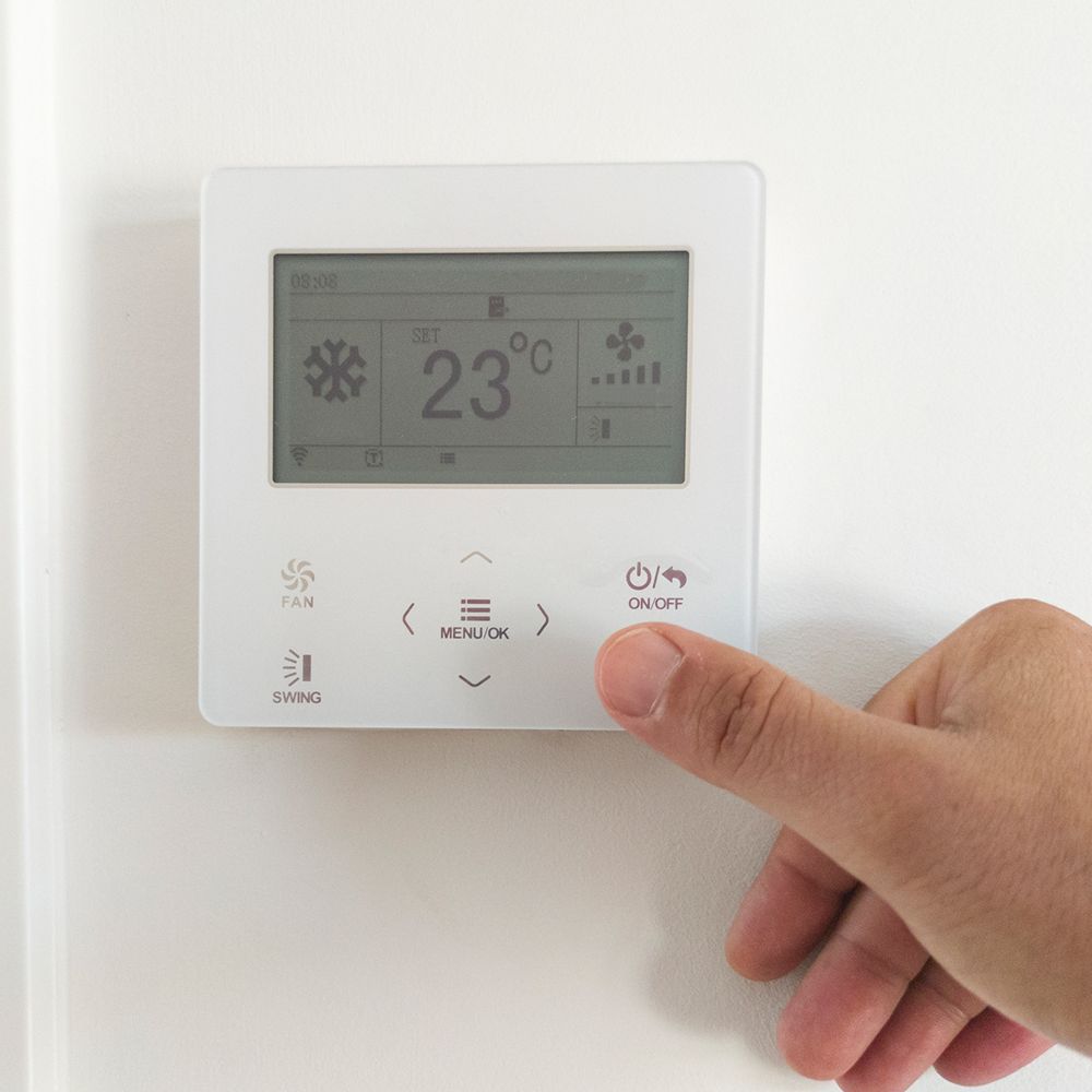 Hand pressing on/off button of a white thermostat displaying 23°C and snowflake icon, mounted on a white wall.
