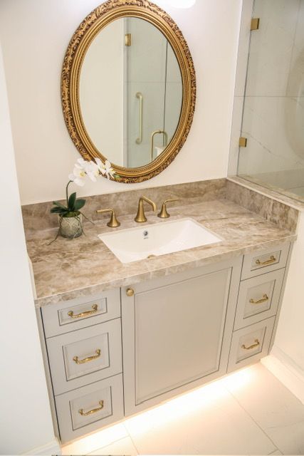 Bathroom vanity with gold accents, oval mirror, gray cabinets, and marble countertop.